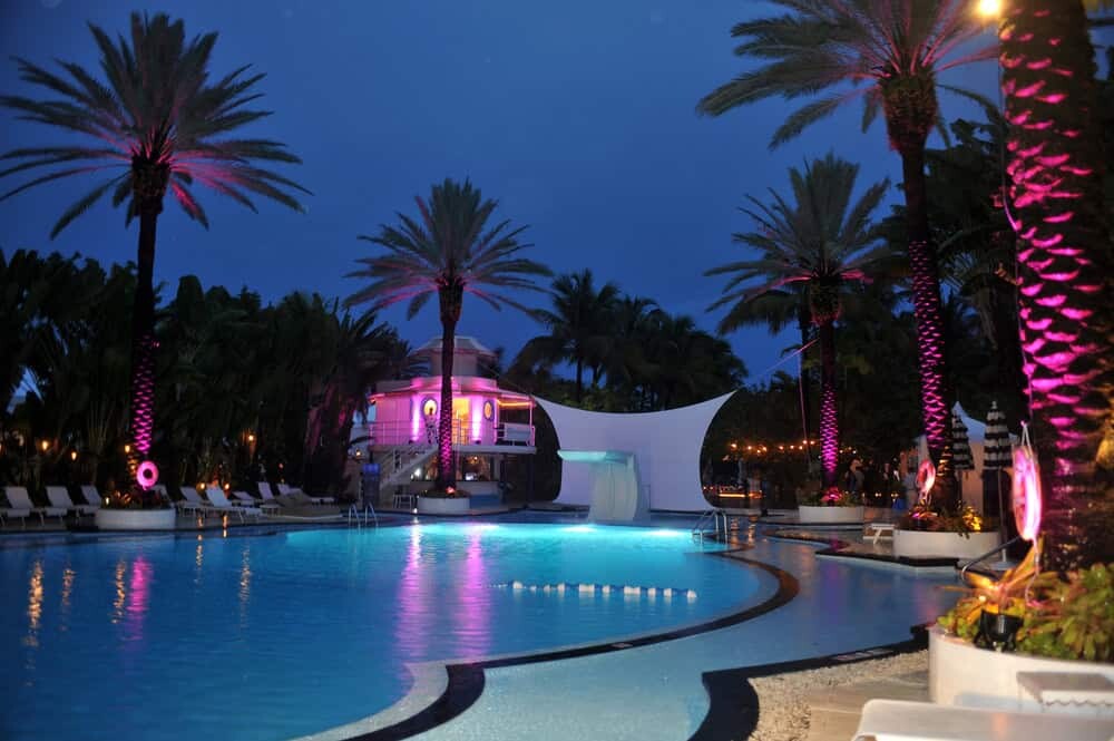 Resort pool at night with illuminated palm trees, a white canopy structure, and lounge chairs surrounding the water-perfect for those seeking upscale swinger vacation destinations in Florida.