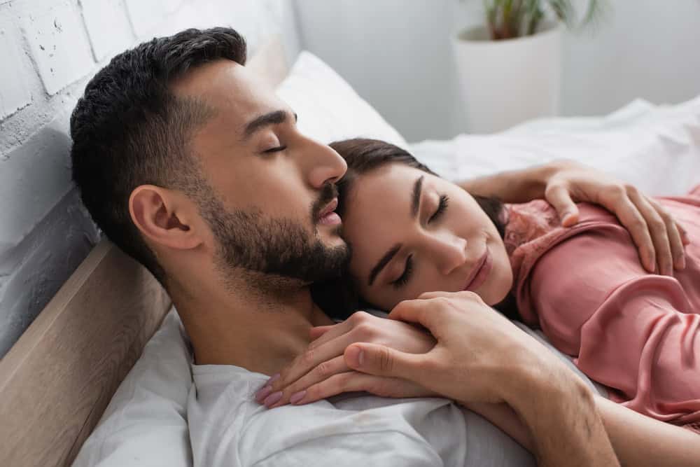A man and a woman lie on a bed, resting together with eyes closed. The woman is nestled against the man's chest, and he holds her hand, sharing an intimate moment that speaks to the closeness of their relationship.