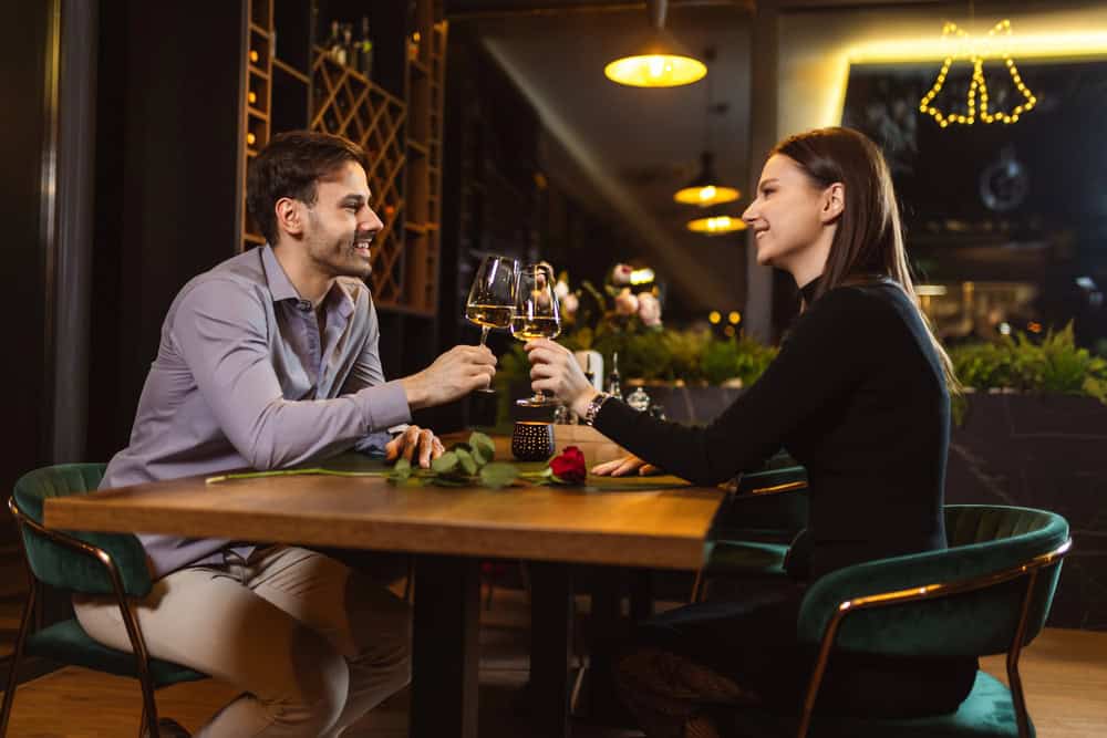 A man and woman sit across from each other at a restaurant table, clinking wine glasses in a sexy, intimate atmosphere, with a single red rose symbolizing their relationship between them.