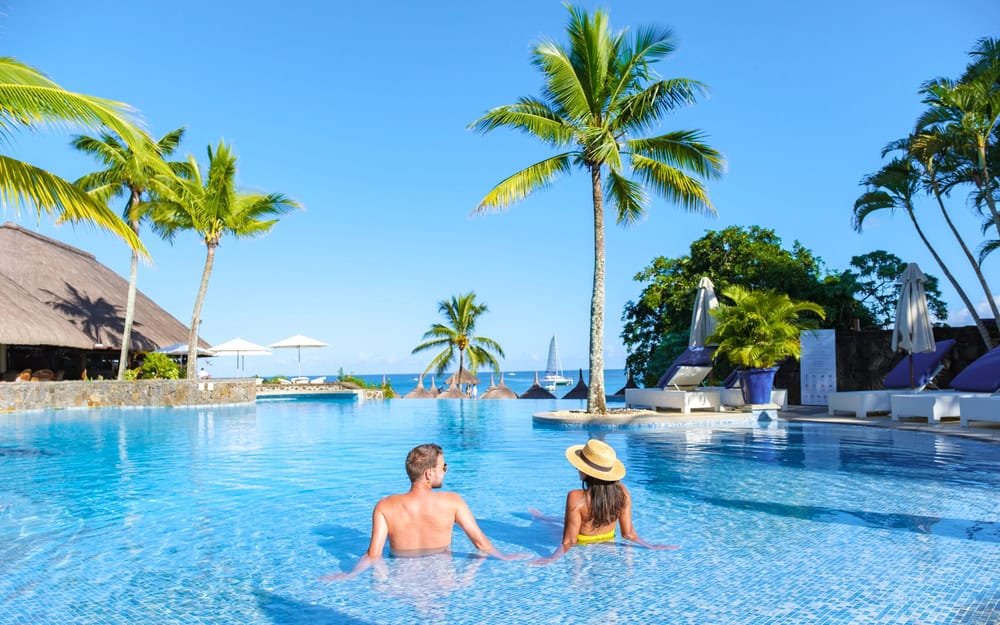 A man and woman relax in a resort pool at one of the best swinger resorts Florida offers, facing the ocean and surrounded by palm trees, lounge chairs, and clear blue sky.